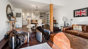 Een moderne woonkamer met een bruine leren bank, een zwarte fauteuil en een houten salontafel. Er is een eethoek met een kroonluchter en een keuken op de achtergrond.