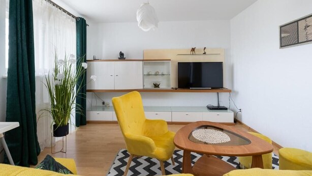 A modern living room with a yellow armchair, a small wooden table, and a TV mounted on a wooden shelf.