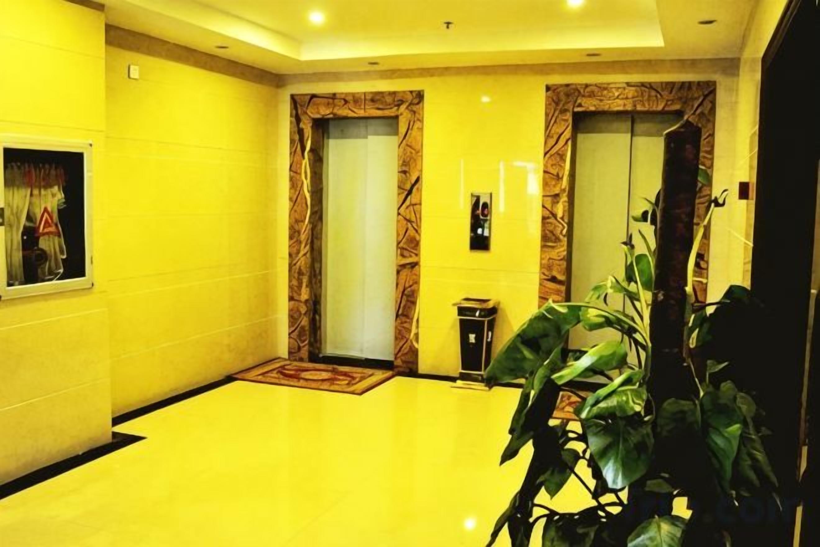 Um saguão de hotel com piso amarelo, dois elevadores, um balcão de recepção e um vaso de planta.