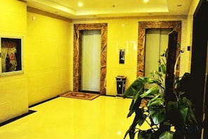 Een hotel lobby met een gele vloer, twee liften, een receptiebalie en een potplant.