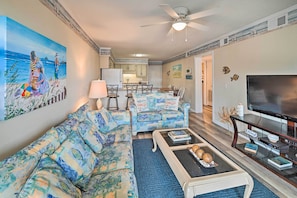 A living room with a sofa, coffee table, and TV. There is a painting on the wall and a ceiling fan.