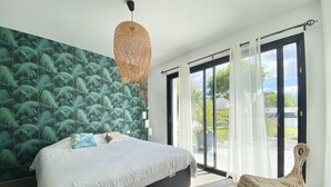 A bedroom with a large bed, a woven chair, a view of the outdoors through a sliding door, and a tropical-themed wall. - Design Villa with Heated Pool, Garden & Beaches 5 min Away (Concarneau)