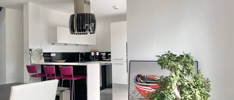 A modern kitchen with white cabinets, a stainless steel range hood, and a dining area with a white chair and a potted plant.