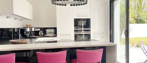 A modern kitchen with white cabinets, a central island with pink bar stools, and a large window with a view of greenery.