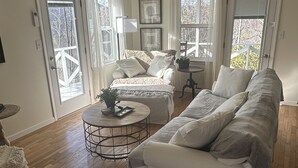 Un salon spacieux avec un grand canapé, une table basse ronde et un tapis à motifs. Il y a deux fenêtres avec des rideaux, une porte et une petite table sur laquelle est posée une plante.