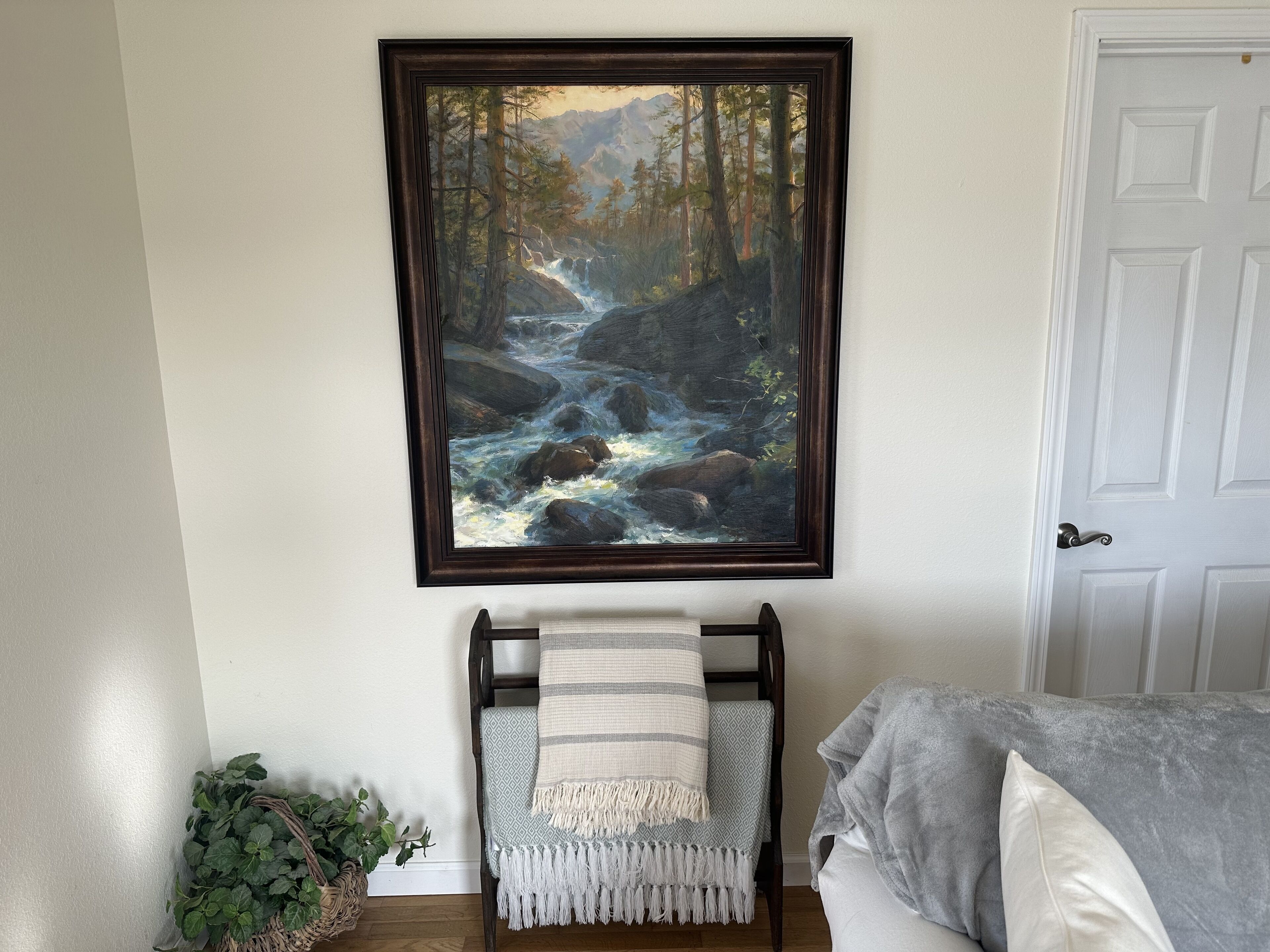 A room with a framed painting of a forest stream, a chair with a patterned throw, and a potted plant.