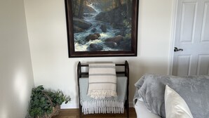 Une pièce décorée d’un tableau encadré représentant un ruisseau de forêt, d’un fauteuil recouvert d’un plaid à motifs et d’une plante en pot.