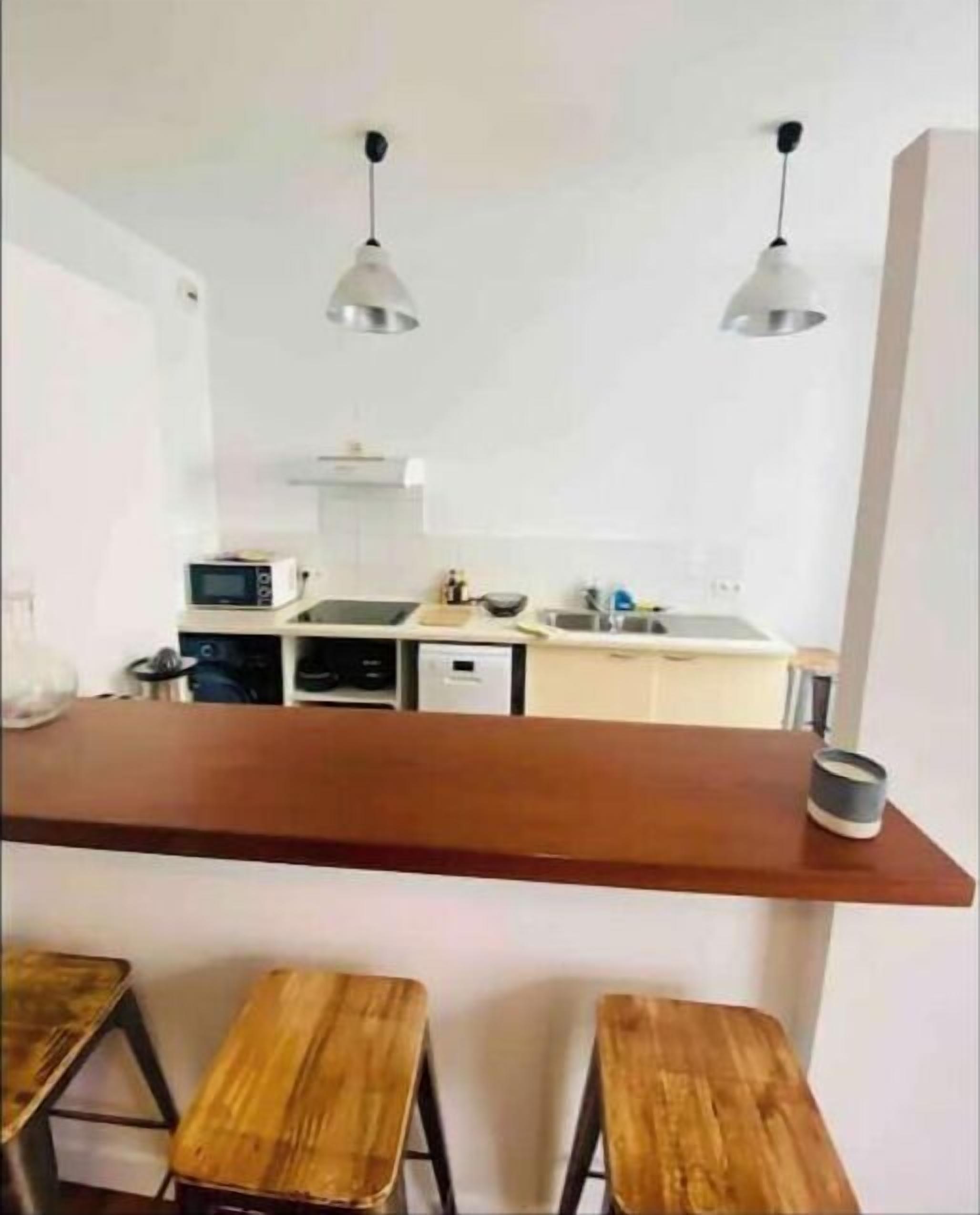Cocina compacta con mostrador y taburetes de madera, equipada con electrodomésticos básicos e iluminación colgante.