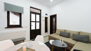 Una sala de estar moderna con un sofá, una mesita redonda y un banco. Hay dos puertas y una ventana con persianas.