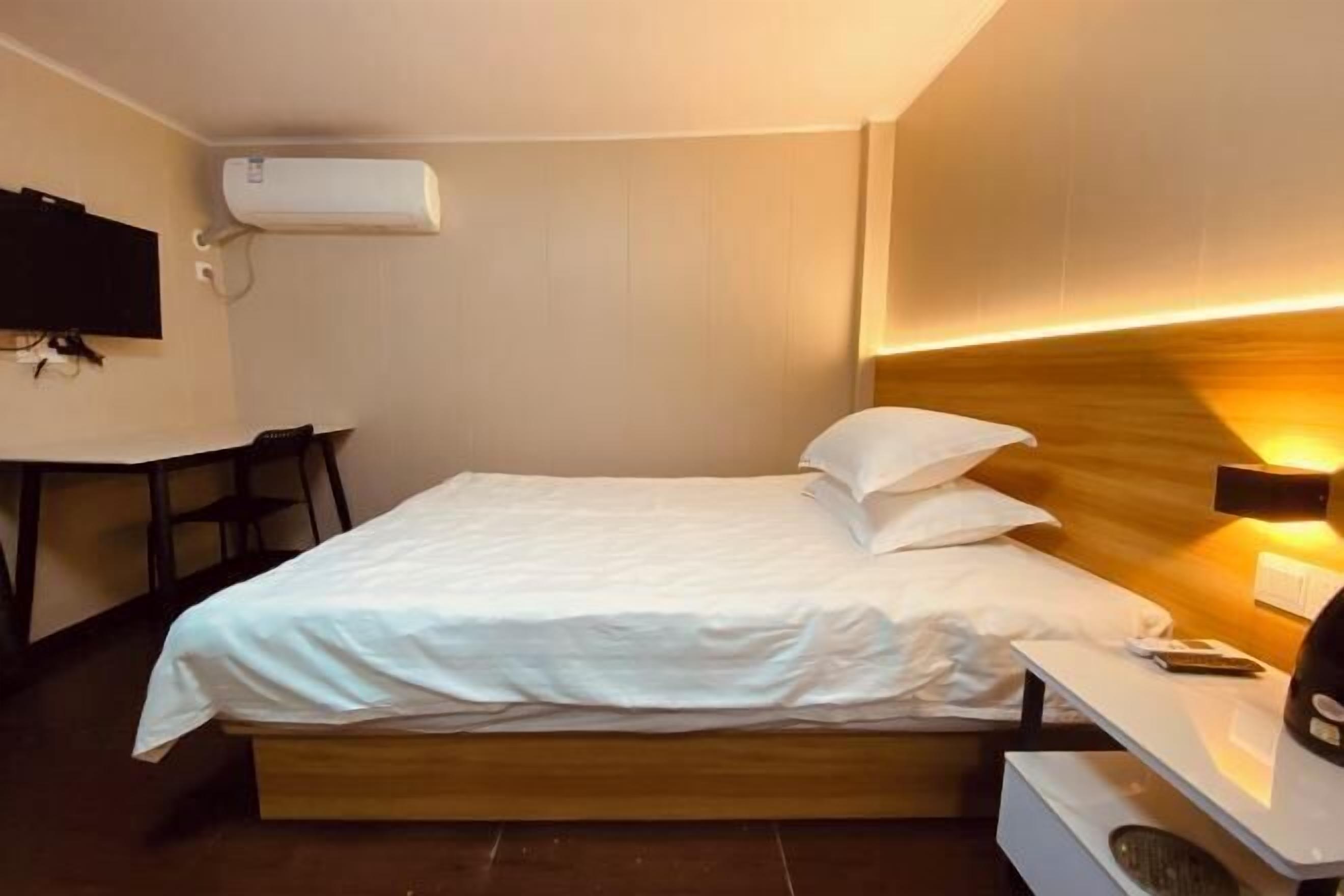 A hotel room with a bed, bedside table, lamp, TV, and air conditioning unit.
