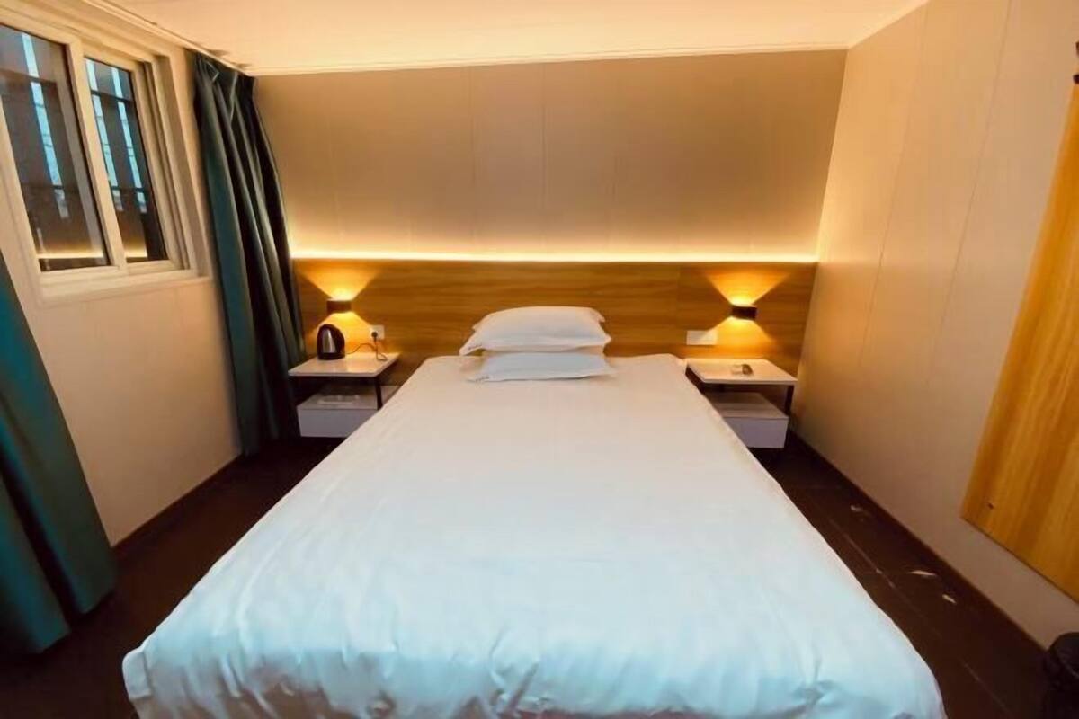 Habitación de hotel con una cama individual, mesitas de noche, cortinas en una ventana y una lámpara fijada a la pared.