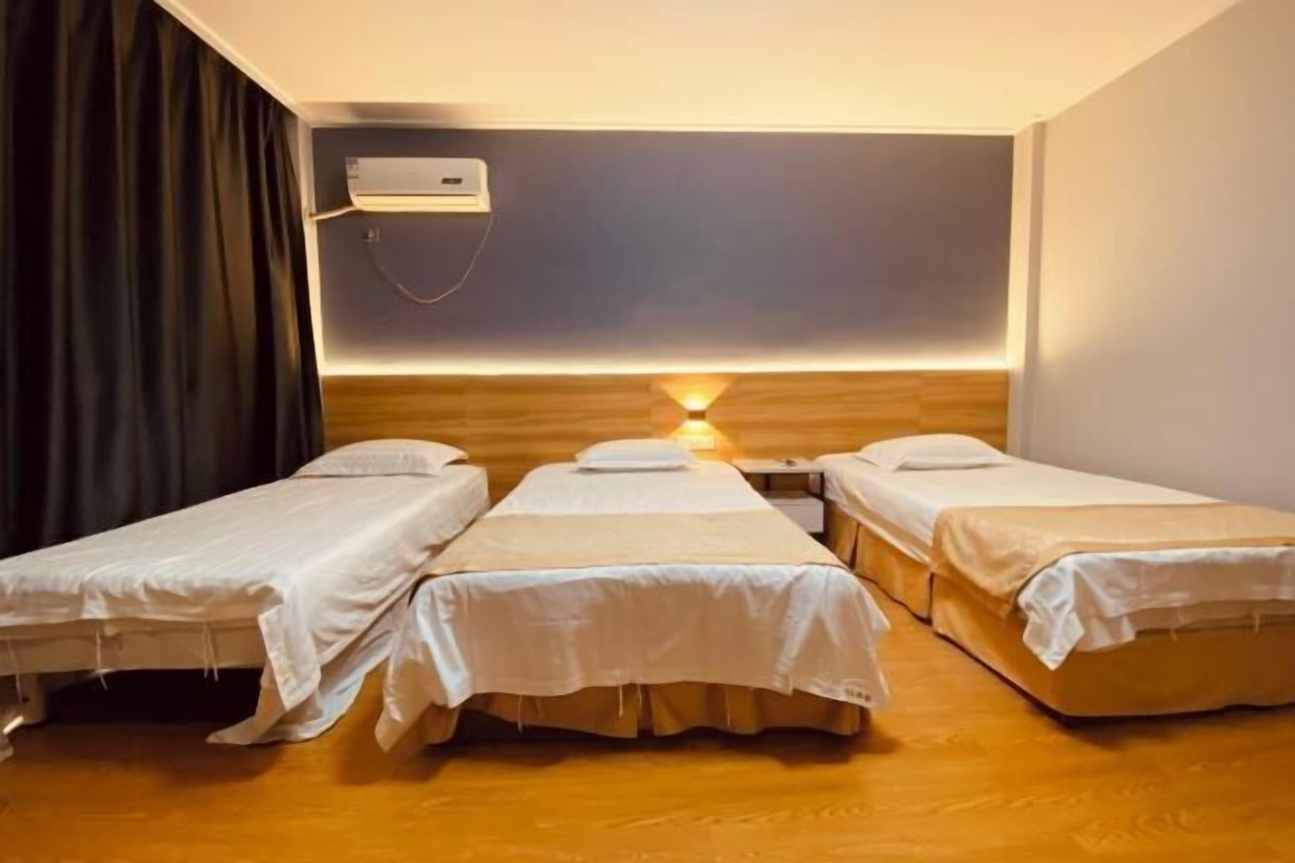 A room with three single beds, a wall-mounted air conditioner, and a bedside table with a lamp.
