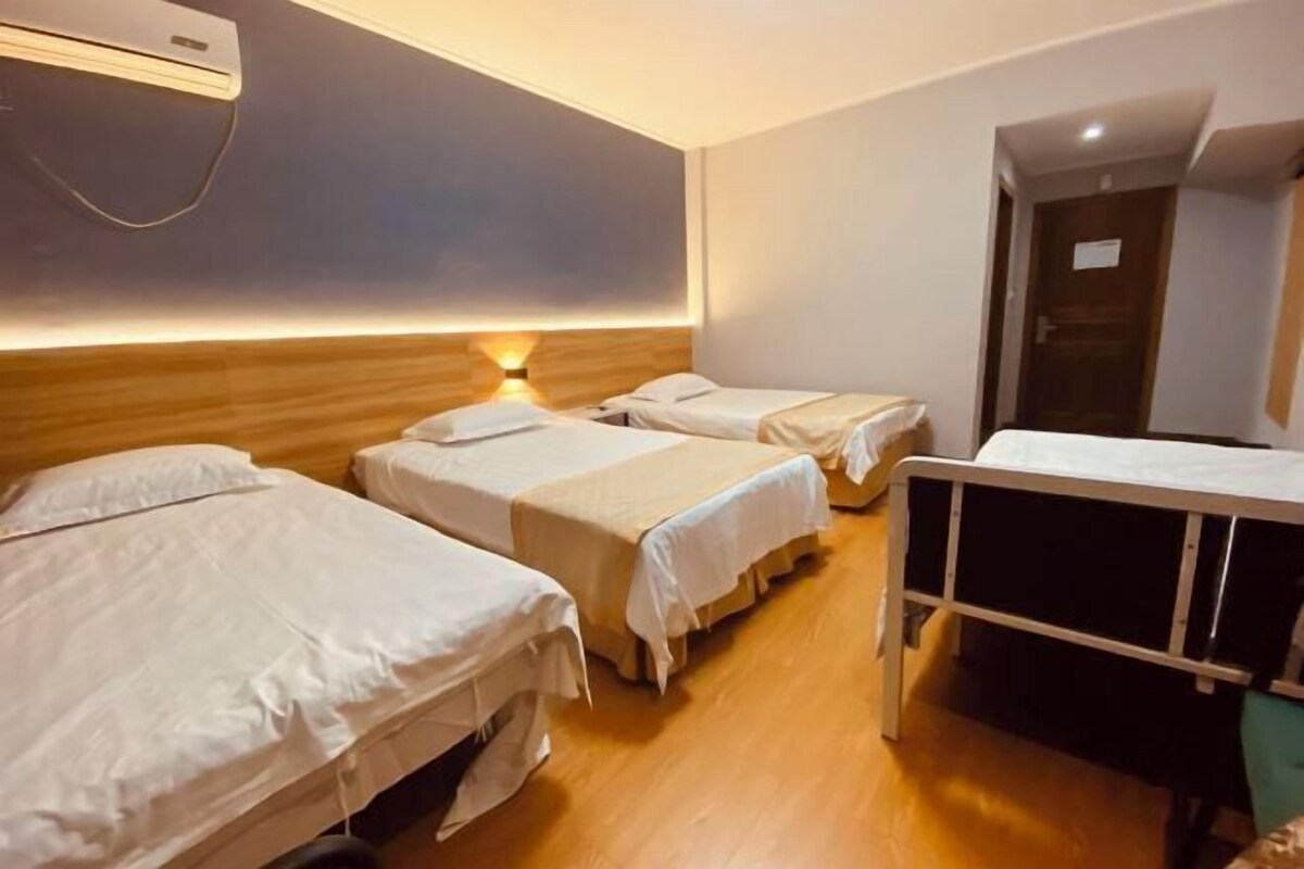 A hotel room with three single beds, a bedside table, a small desk, and a wall-mounted air conditioner.