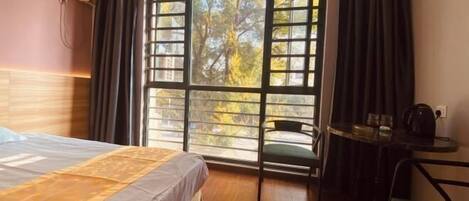A hotel room with a bed, a small table with a kettle, a chair, and a window with a view of trees.