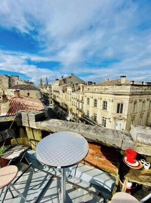 A rooftop terrace with a round table and chairs, overlooking a cityscape with historic buildings.