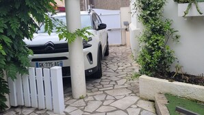 Un camino adoquinado de piedra conduce a un coche blanco estacionado bajo un arco blanco, rodeado de follaje y una cerca blanca.