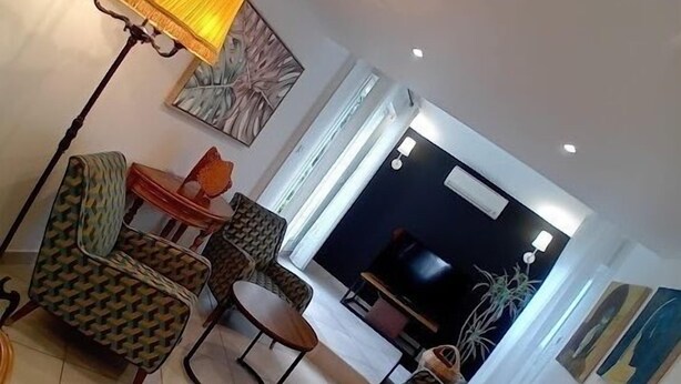 Una sala de estar con sofás estampados, una mesa de centro y una lámpara de pie. Se observa un televisor y una planta al fondo.