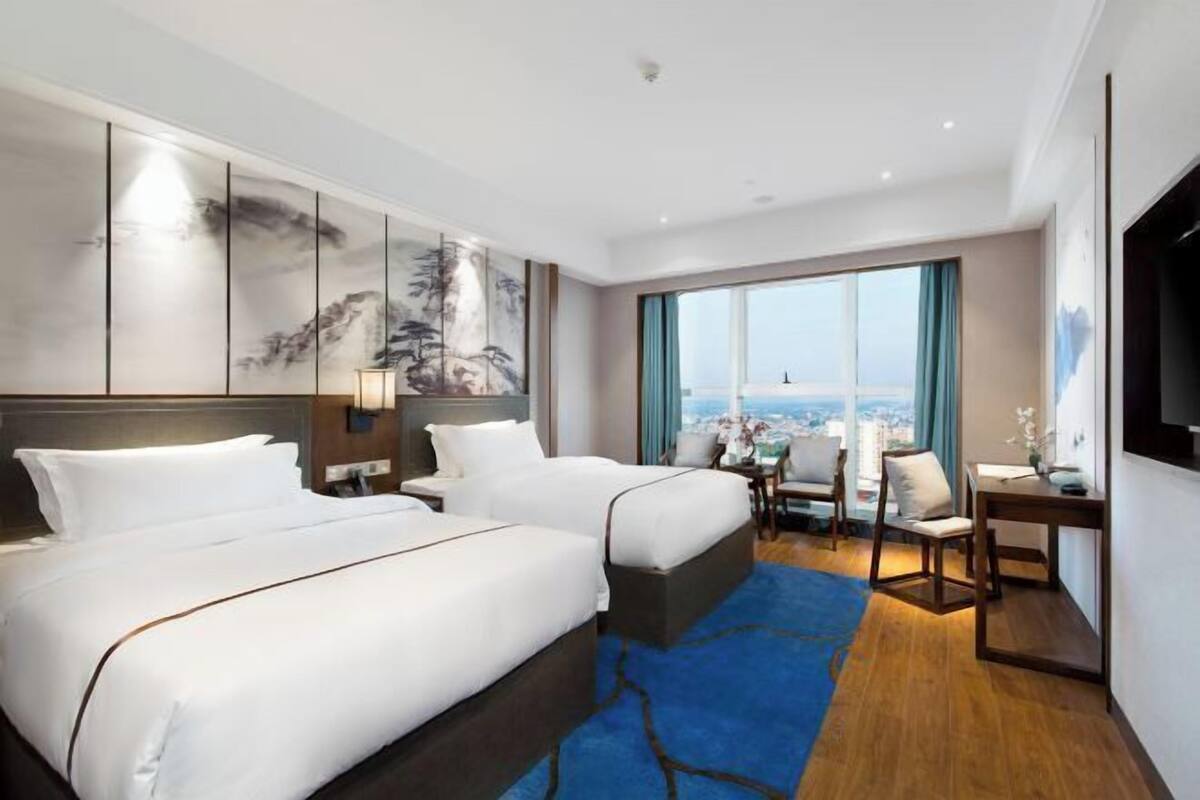 A hotel room with two beds, a desk, a chair, and a large window with a city view.