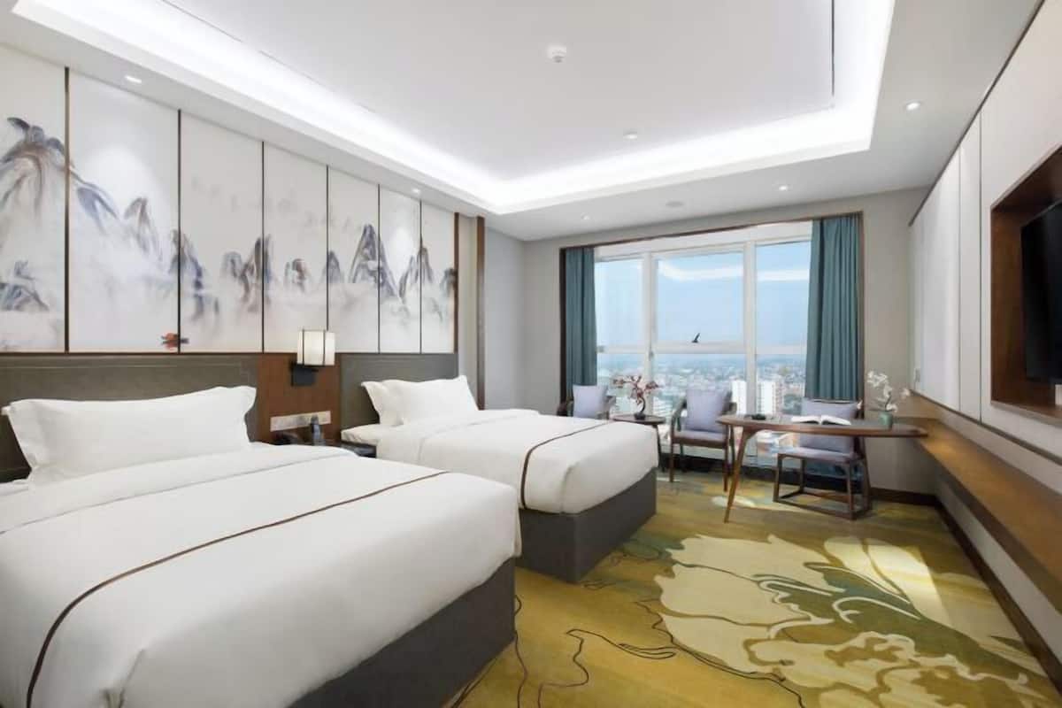 A hotel room with two beds, a large window with a city view, a dining area, and a painting on the wall.