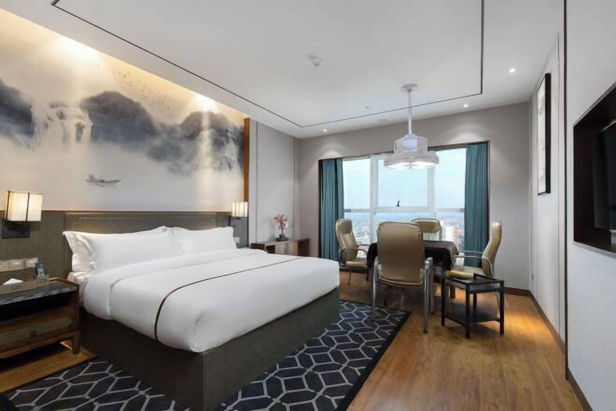 A modern hotel room with a large bed, a desk with two chairs, a view of the city through large windows, and a painting on the wall.