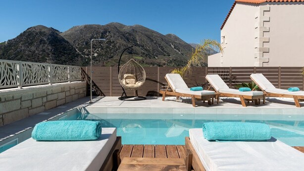 Un espace piscine doté de chaises longues et d’un fauteuil suspendu, entouré d’une terrasse en bois et d’une clôture blanche, avec des montagnes en arrière-plan.