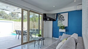 A modern living room with a sofa, a small round table, and a glass door leading to an outdoor area with a pool and patio furniture.
