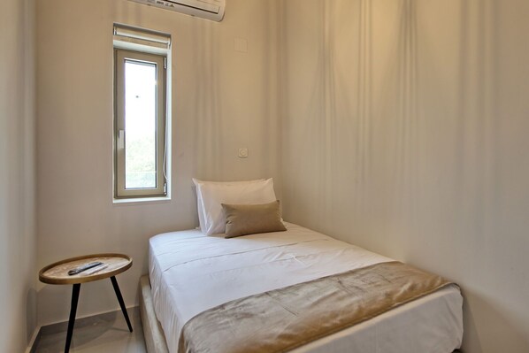 Una cama pequeña y ordenada en una habitación con un ventanal, una mesita redonda y una pared sencilla.