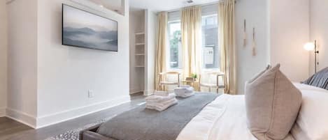 Un hotel moderno con cama, televisor montado en la pared, una pequeña zona de estar con sillones y vistas al exterior a través de amplios ventanales.