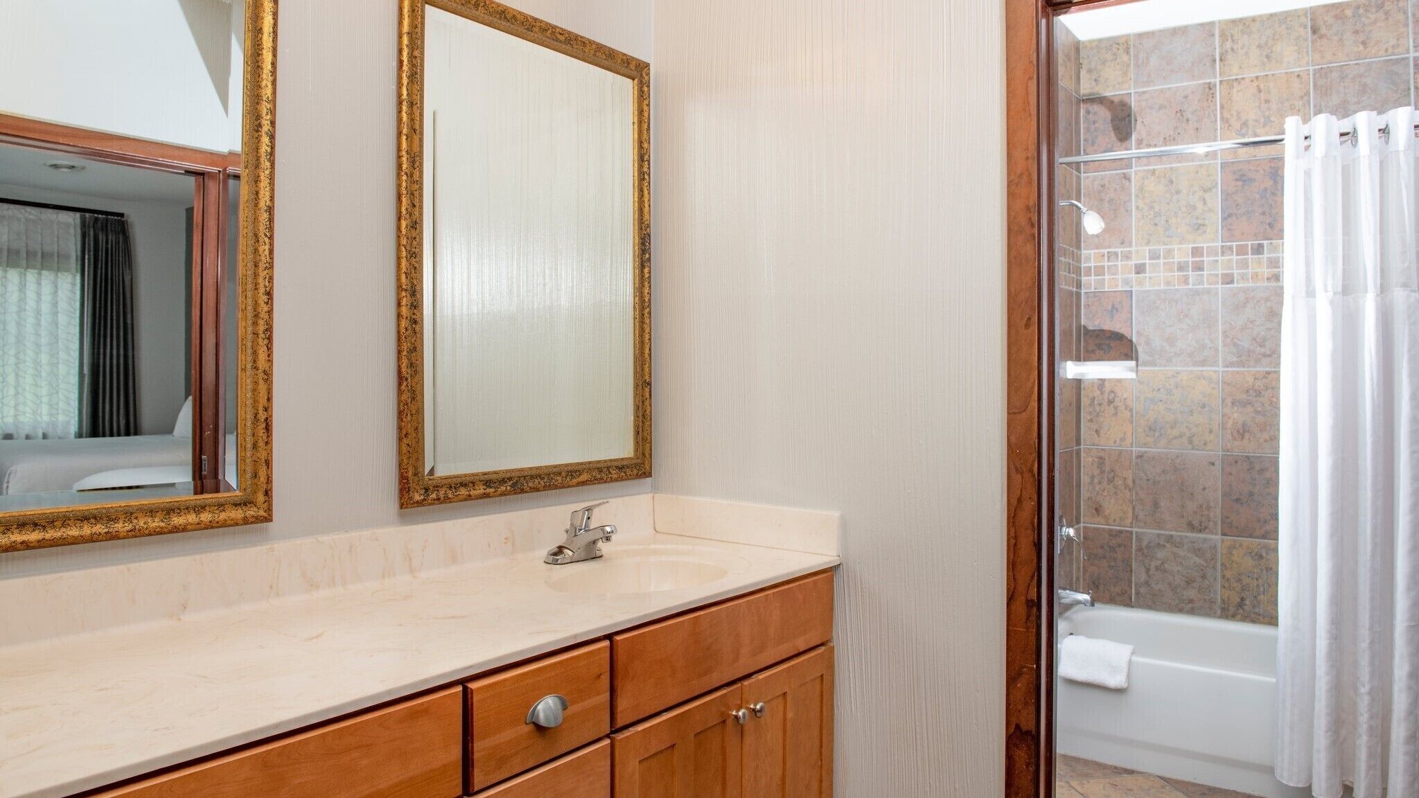 Une salle de bain avec un lavabo en marbre, un meuble en bois, deux miroirs avec des cadres dorés et un espace douche avec un mur carrelé et un rideau blanc.