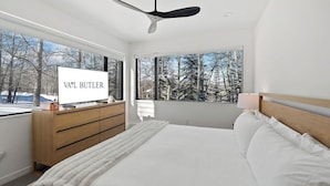 Een moderne slaapkamer met een bed, een televisie op een houten dressoir en grote ramen met uitzicht op bomen.