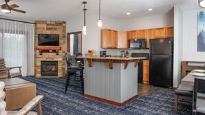 Modern kitchen with central island, microwave, refrigerator, stone fireplace, and television.