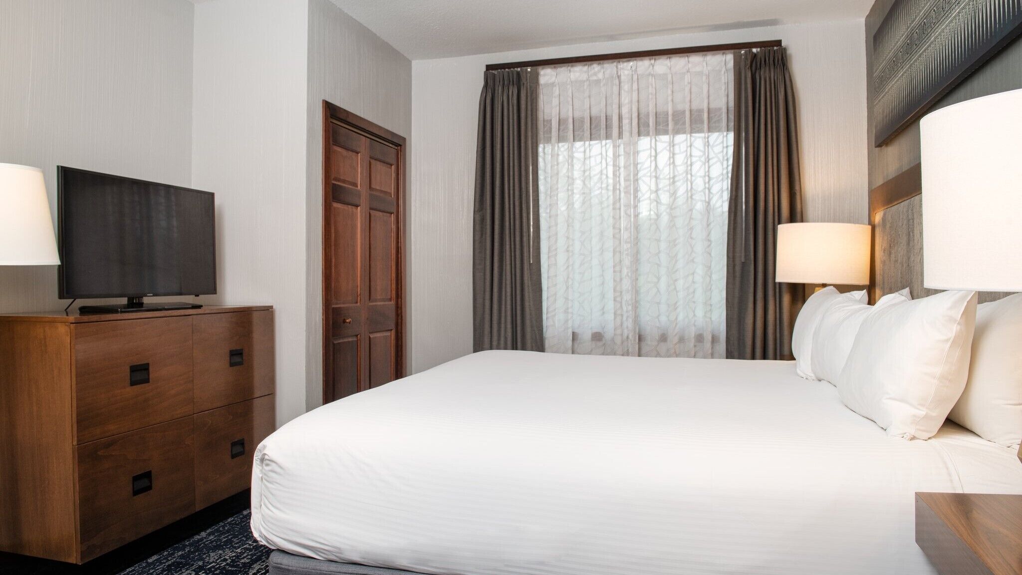 A hotel room with a large bed, a TV on a wooden dresser, a wardrobe, and a window with curtains.