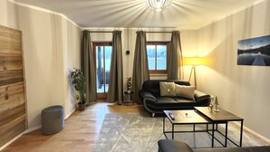 A modern living room with a sofa, a coffee table, and a lamp. There is a view through the window, a plant, and a wooden wall. - Spacious apartment with sun terrace in the immediate vicinity of the lake (Bad Wiessee)