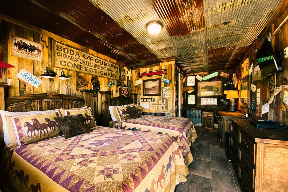 A rustic room with wooden walls and a ceiling, featuring a bed with a quilted horse pattern, a wooden bench, and various signs including one for a sodas fountain and a no parking sign.