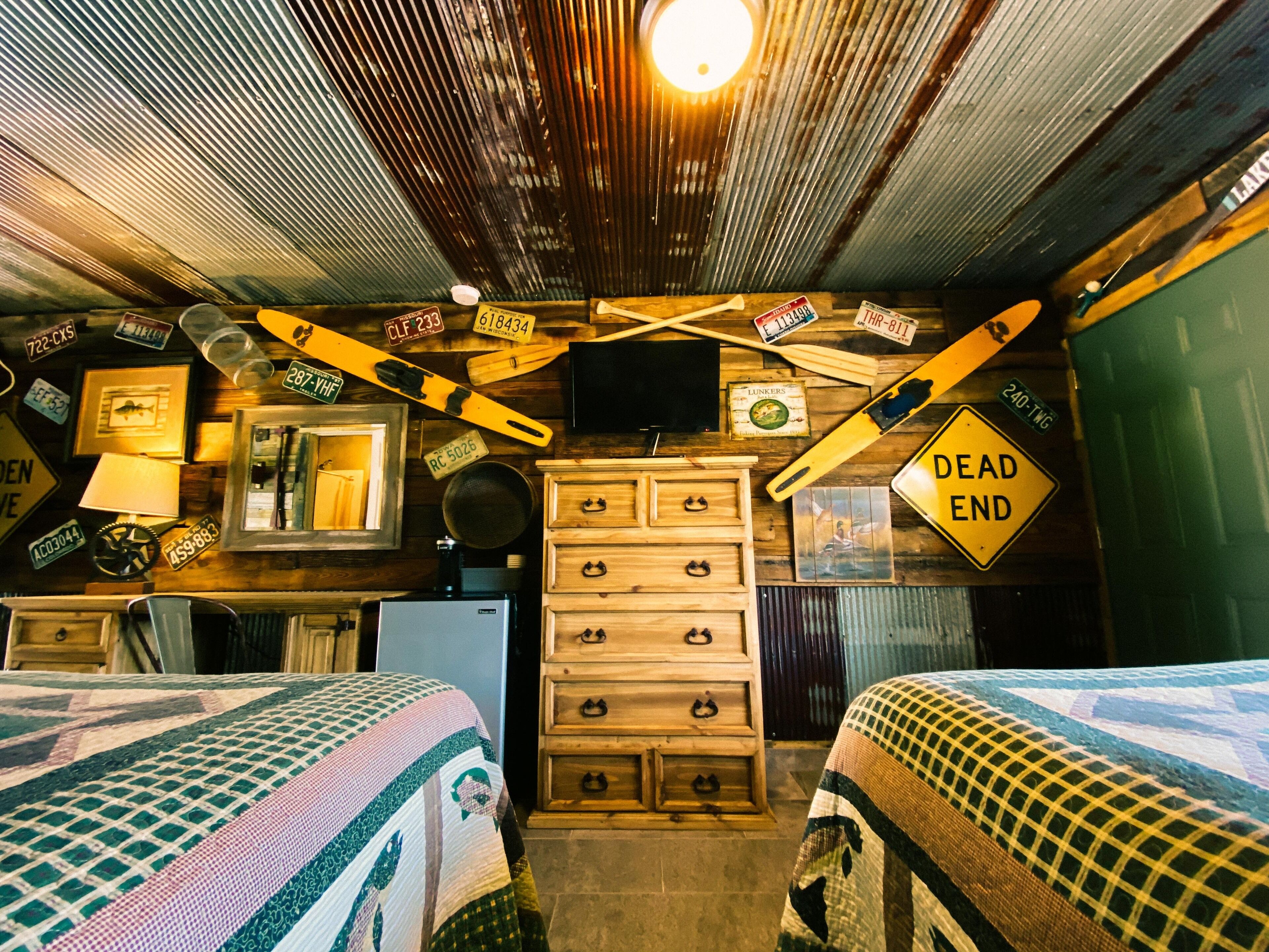 A room with two beds, a dresser, a television, and various decorative items including license plates and a 'Dead End' sign.