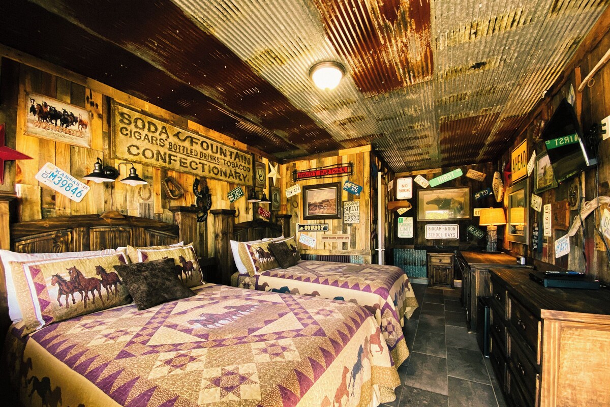 Ein rustikaler Raum mit Holzwänden und -decken, der ein Bett mit einem gepolsterten Pferdemuster, eine hölzerne Bank und verschiedene Schilder, darunter eines für eine Limonadenbar und ein Verbotsschild, zeigt.