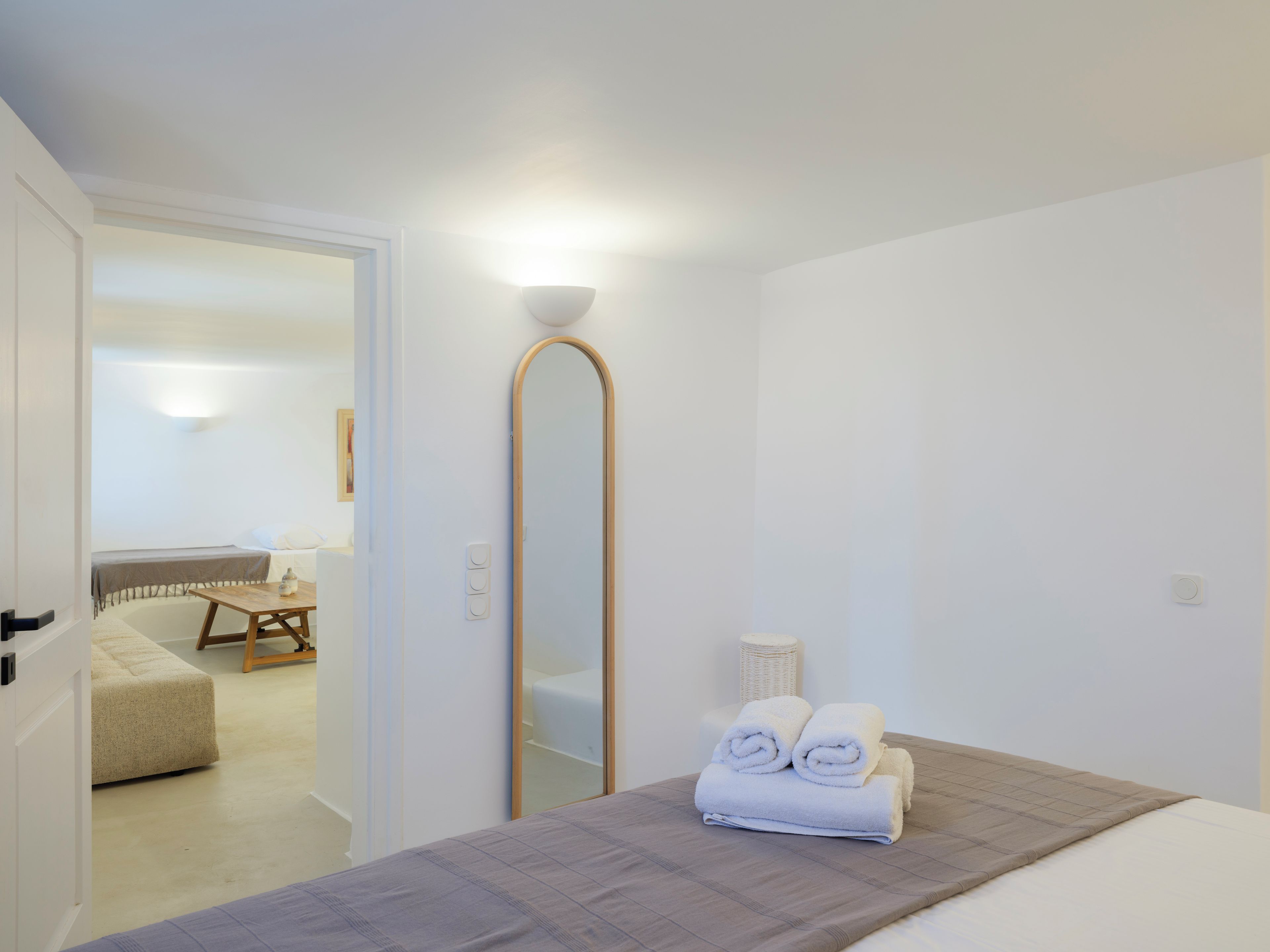 A hotel room with a bed, a mirror, a bedside table, and a view into the adjacent room.