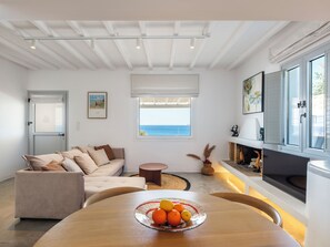 Uma sala de estar moderna com um sofá, uma mesa de jantar com frutas, uma lareira e vista para o mar.