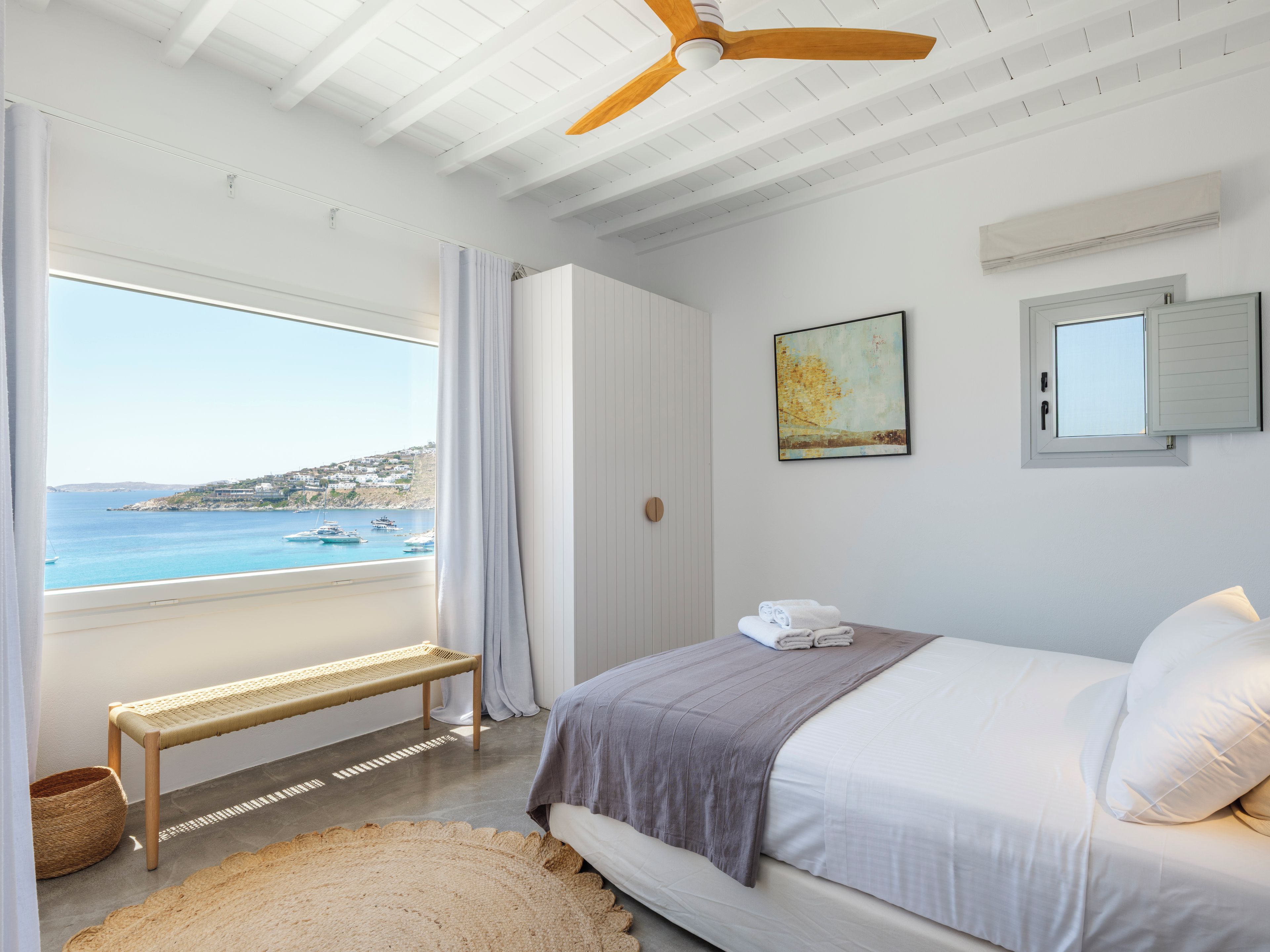 Een slaapkamer met een bed, een bankje, uitzicht op zee en een plafondventilator.