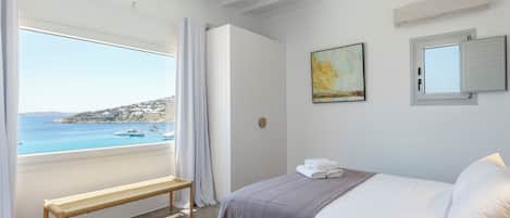 Een slaapkamer met een bed, een bankje, uitzicht op zee en een plafondventilator.
