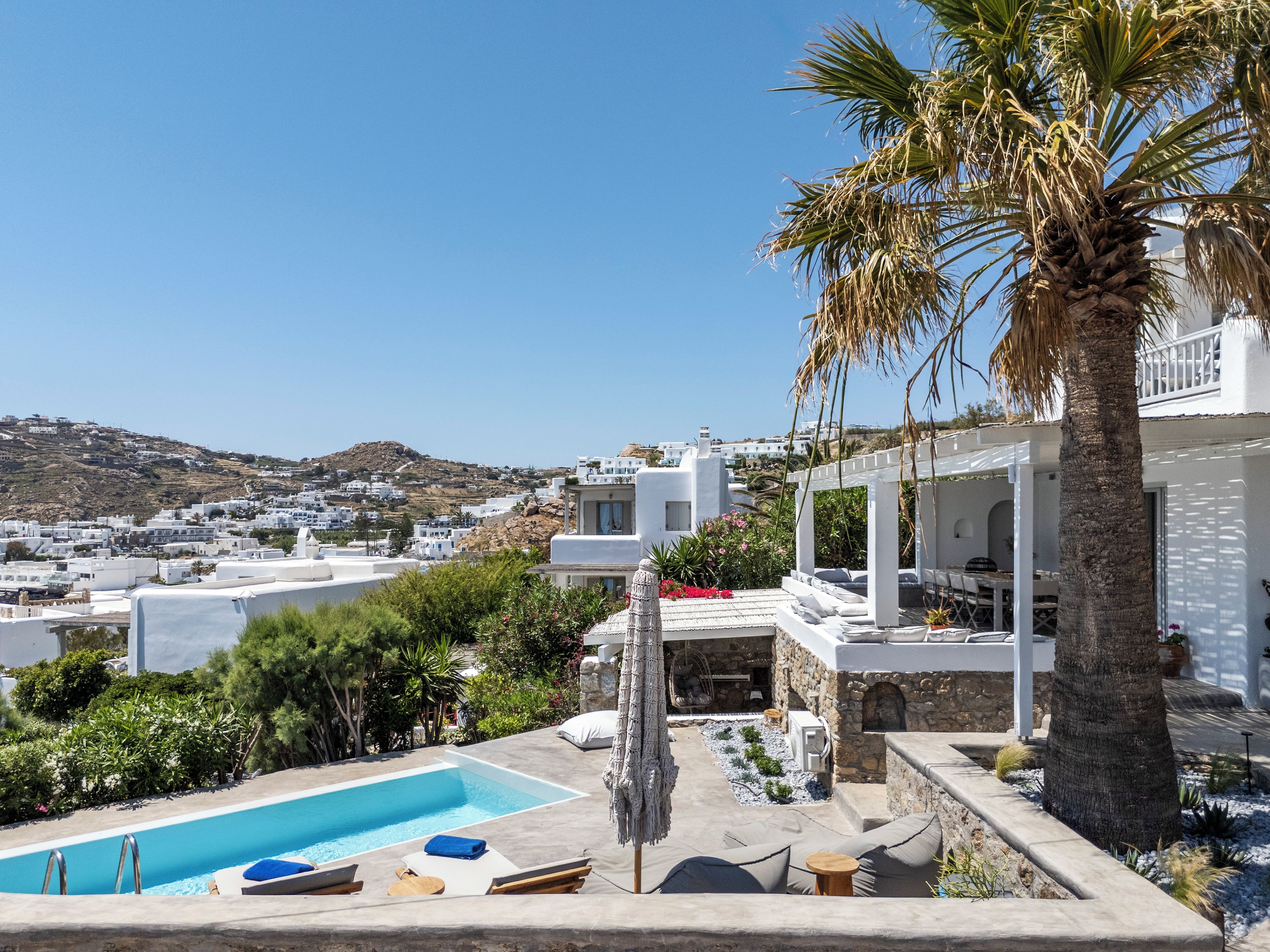 Zona de piscina con sillones de descanso y una pared de piedra, con vistas a un pueblo costero de edificios blancos.