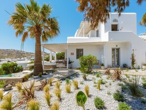 A white building with a covered patio area, surrounded by various plants and a palm tree. - MyMykonos Villa II Beach Front  (Mykonos)