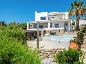 Um edifício moderno de dois andares com fachada de pedra, piscina e vegetação exuberante.