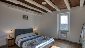 A bedroom with a wooden ceiling, a large bed with a grey headboard, two bedside tables with lamps, a window with a view, and a small picture on the wall. - La Petite Ferme near DOMME8FP3Q6W9+8R 44.7958121,1.2195112 (Domme)