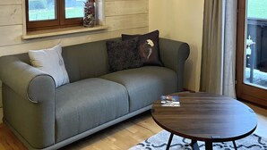 A living room with a grey sofa, a round wooden coffee table, and a window with curtains. - In the immediate vicinity of the lake, small, fine apartment newly renovated (Bad Wiessee)