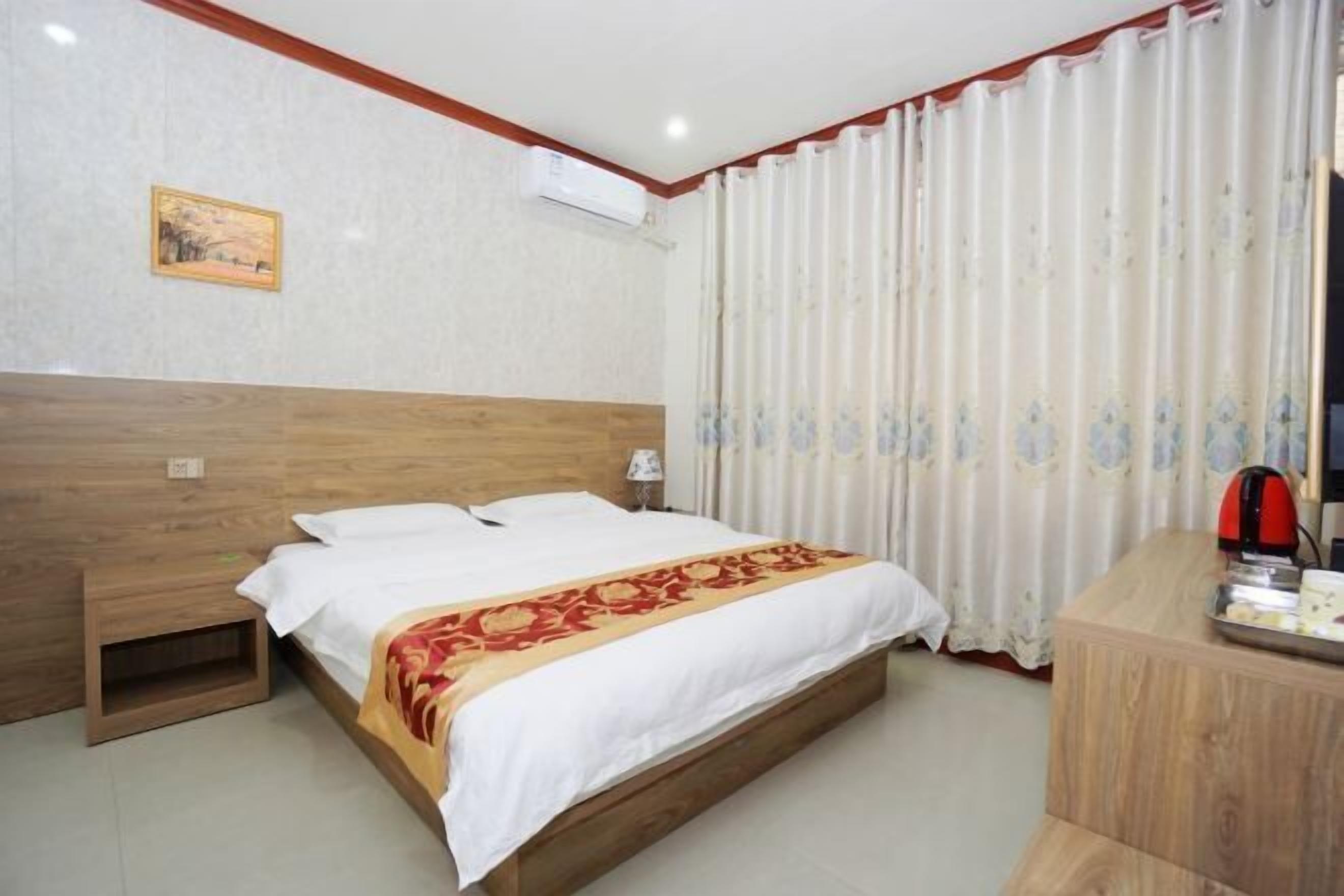 Habitación de hotel con cabecera de madera, cama con ropa blanca y una manta roja y dorada, mesita de noche de madera, escritorio con hervidor y ventana con cortinas blancas.