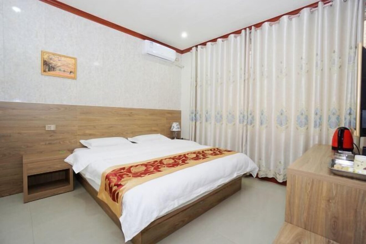 Ein Hotelzimmer mit hölzernem Kopfteil, einem Bett mit weißer Bettwäsche und einer roten und goldenen Tagesdecke, einem Nachttisch aus Holz, einem Schreibtisch mit Wasserkocher und einem Fenster mit weißen Vorhängen.