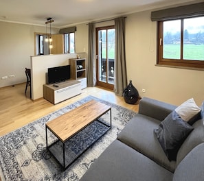 A modern living room with a sofa, coffee table, TV, and a view of a green landscape through large windows.
