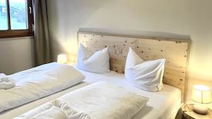 Una cama doble con sábanas y almohadas blancas, un cabecero de madera, una mesita de noche con una lámpara y una ventana con cortinas.