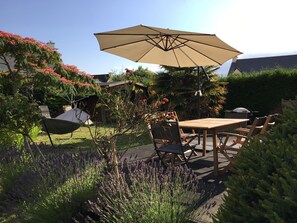 Eine Terrasse mit Holztisch und -stühlen, einem großen Sonnenschirm und einem Garten mit Lavendel und blühenden Sträuchern.